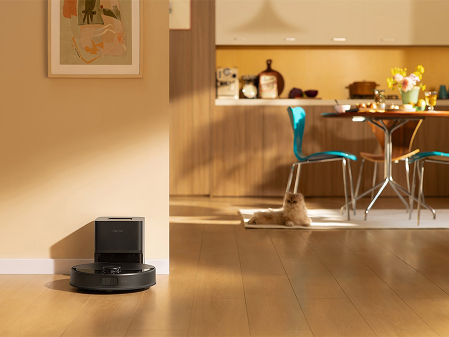 Saugroboter Roborock Q10 reinigt den Boden in der Nähe einer Katze auf dem Teppich; im Hintergrund gedeckter Esstisch