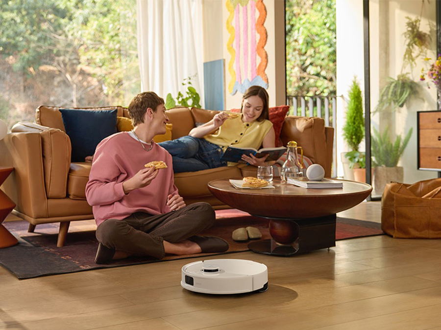 Zwei junge Menschen entspannen im Wohnzimmer, während der weiße Staubsaugerroboter Roborock Q10 PF White den Boden saugt