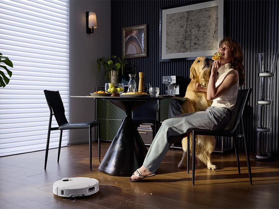 Frau isst Pizza am Esstisch mit einem Hund, während der Saugroboter Roborock Qrevo Edge T Weiß Krümel vom Boden aufsaugt