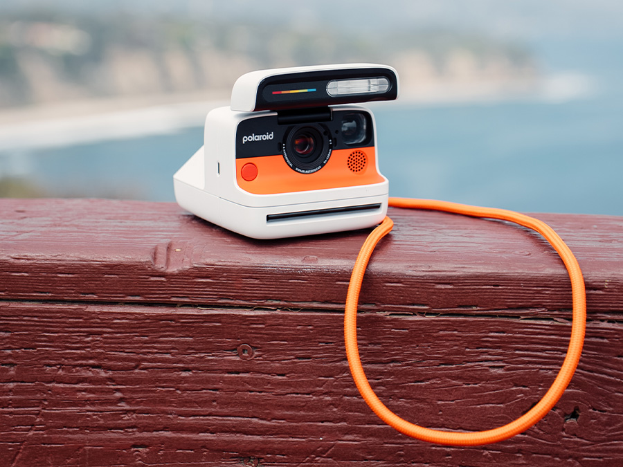 Weiße Polaroid Flip Sofortbildkamera mit orangefarbenem Front und Tragegurt, liegend auf einer Holzbrüstung mit unscharfem Meereshintergrund