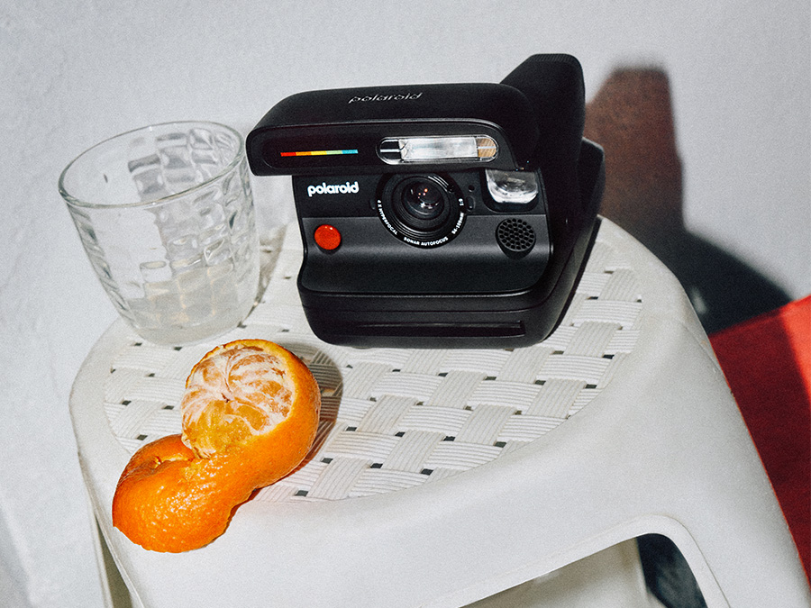 Schwarze Sofortbildkamera Polaroid Flip auf weißem, geflochtenem Hocker neben einem Glas und einer geschälten Mandarine