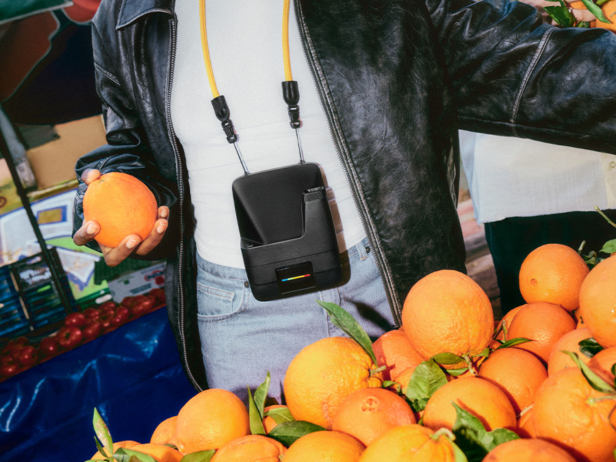 Person in schwarzer Lederjacke hält eine Orange auf einem Markt, mit schwarzer Sofortbildkamera Polaroid Flip, die an einem gelben Band um den Hals hängt
