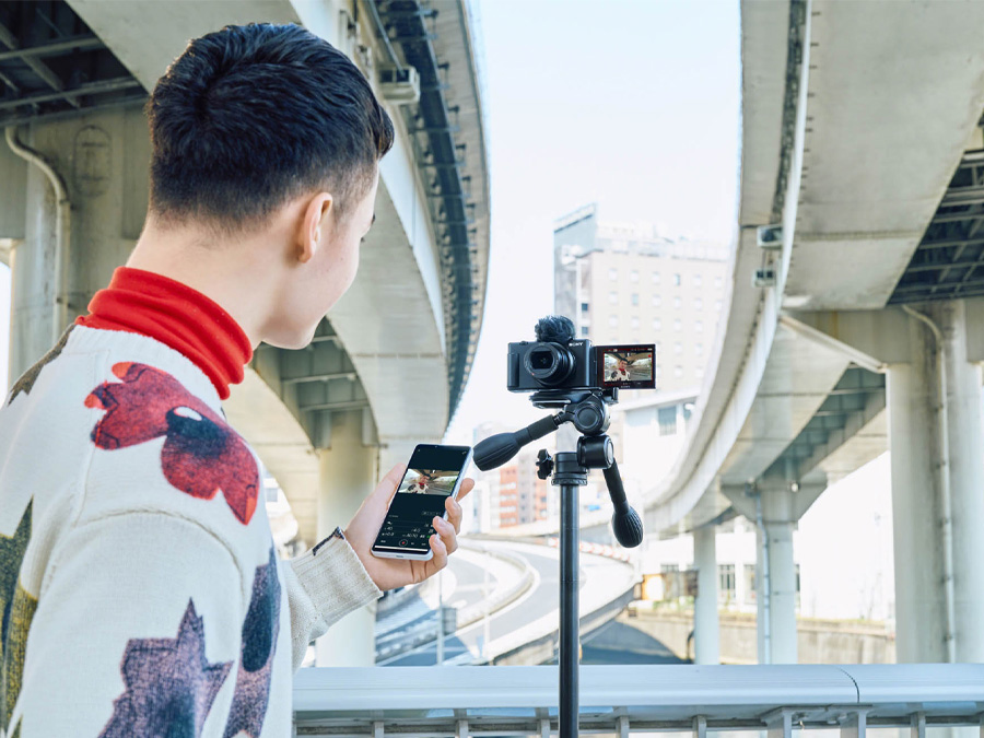 Person filmt mit der Sony ZV-1 II Kamera auf einem Stativ, steuert die Aufnahme mit einem Smartphone
